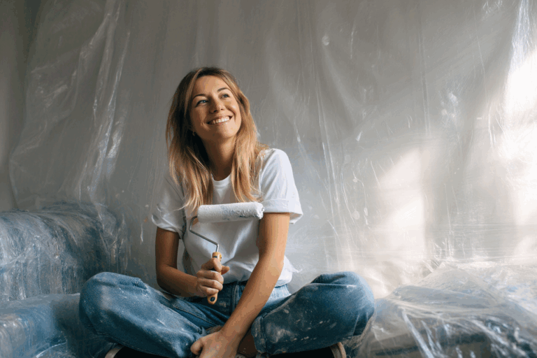 Femme souriante assise sur un canapé protégé par une bâche transparente, rouleau de peinture à la main, mur fraîchement peint en arrière-plan